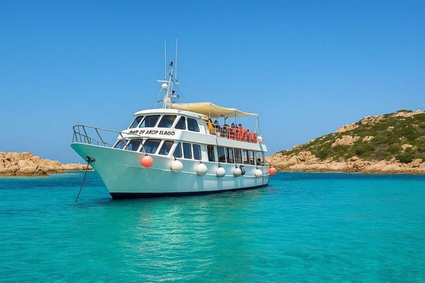 Ein weißes Boot mit der Aufschrift 'GMB OF ARCP ELAGO' schwimmt auf klarem türkisblauem Wasser vor einer felsigen Küstenlinie. 1