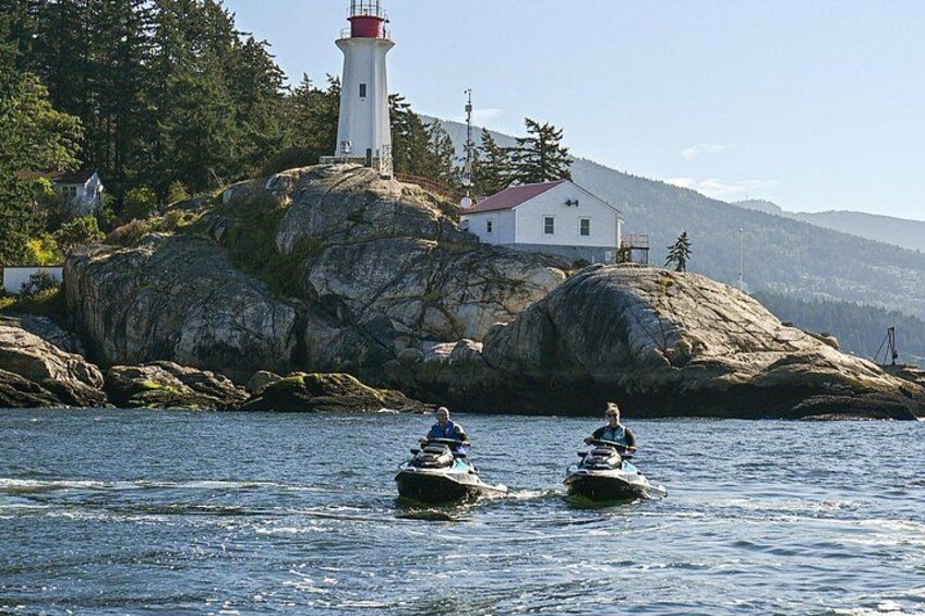 Two people riding jet skis on a body of water near a lighthouse and a white building on a rocky island. 2