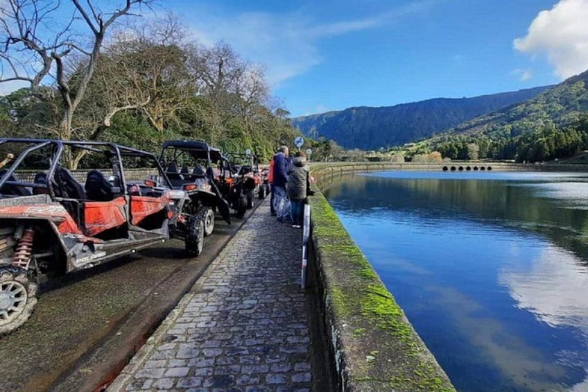 A row of ATVs parked by a calm river, with a bridge in the background and mountains in the distance. 5
