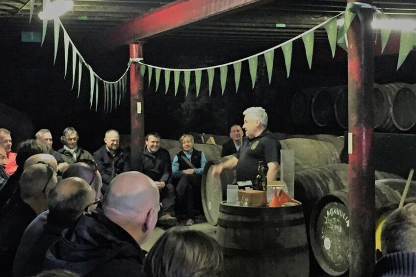 A group of people gathered around a large barrel in an industrial setting with metal structures and pipes in the background. 1