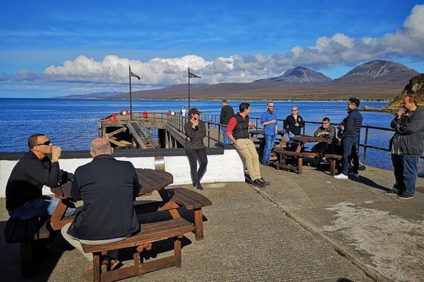 A group of people sitting and standing on a pier overlooking a body of water with mountains in the background. 3