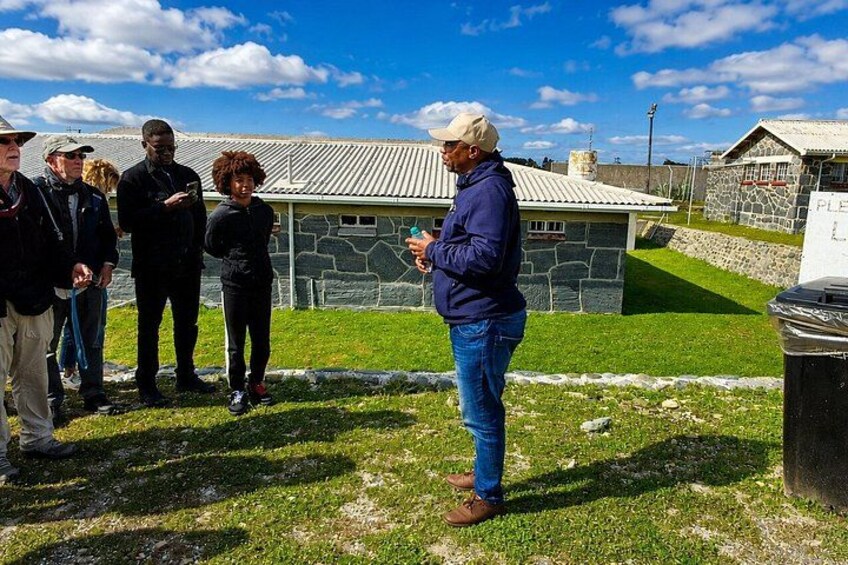 A group of people standing outdoors near stone buildings with a grassy area in the foreground. 2