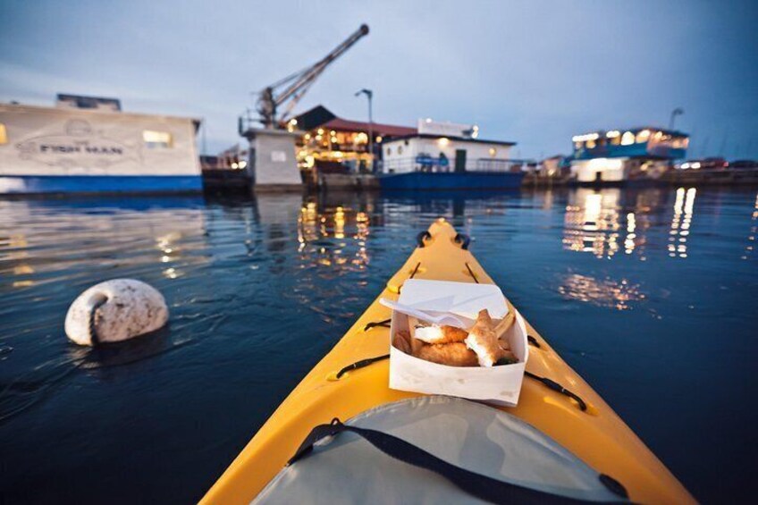 Ein Kajak mit einem Korb voller Proviant auf ruhigem Gewässer, ein Steg und ein großes Schiff im Hintergrund. 4
