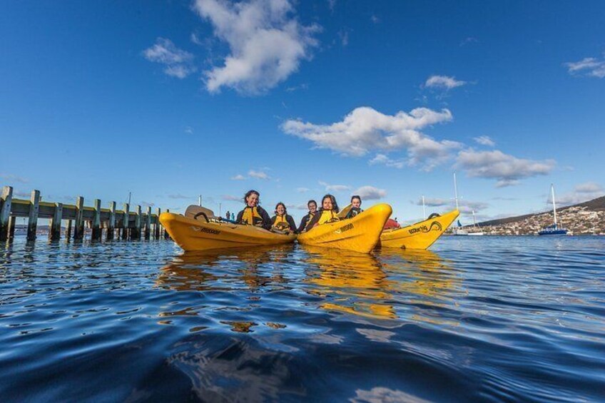 Un groupe de personnes en kayaks jaunes sur une étendue d’eau calme, sous un ciel dégagé, avec une jetée en bois en arrière-plan. 1