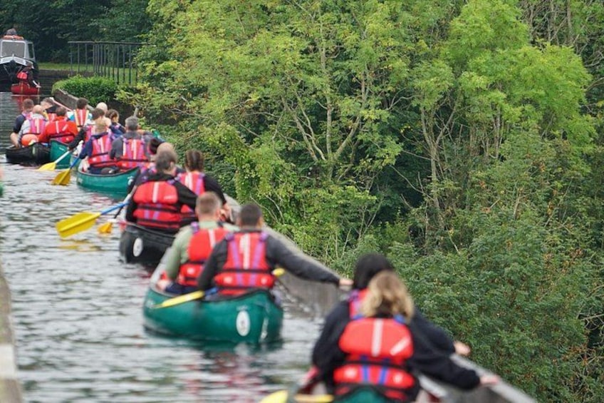 A group of people in green canoes wearing red life jackets, paddling on a river surrounded by dense greenery. 4
