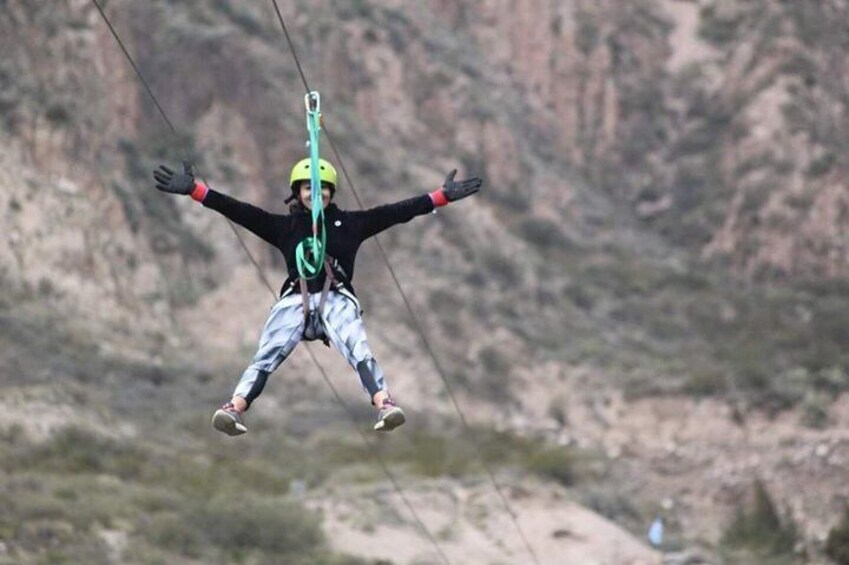 A person paragliding with arms outstretched, wearing a helmet and harness, against a mountainous backdrop. 1