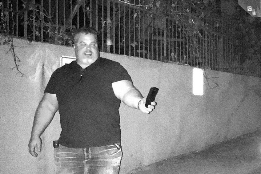 En person holder en smartphone, iført en mørk polo og jeans, står op ad en væg med et metalhegn i baggrunden. 3