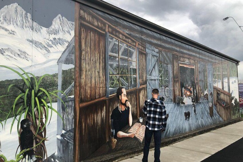 A mural of a rustic wooden house with a mountain backdrop, a person sitting on a bench, and another person standing beside the mural. 4
