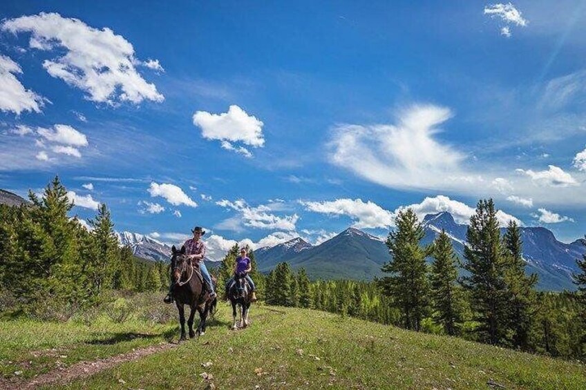 Dos personas cabalgando por un sendero cubierto de hierba, con montañas al fondo. 5