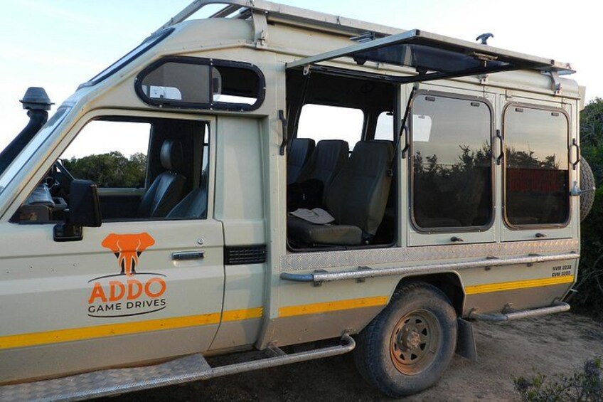 A beige off-road vehicle with 'ADDO GAME DRIVES' branding, equipped with a roof rack and side steps. 2