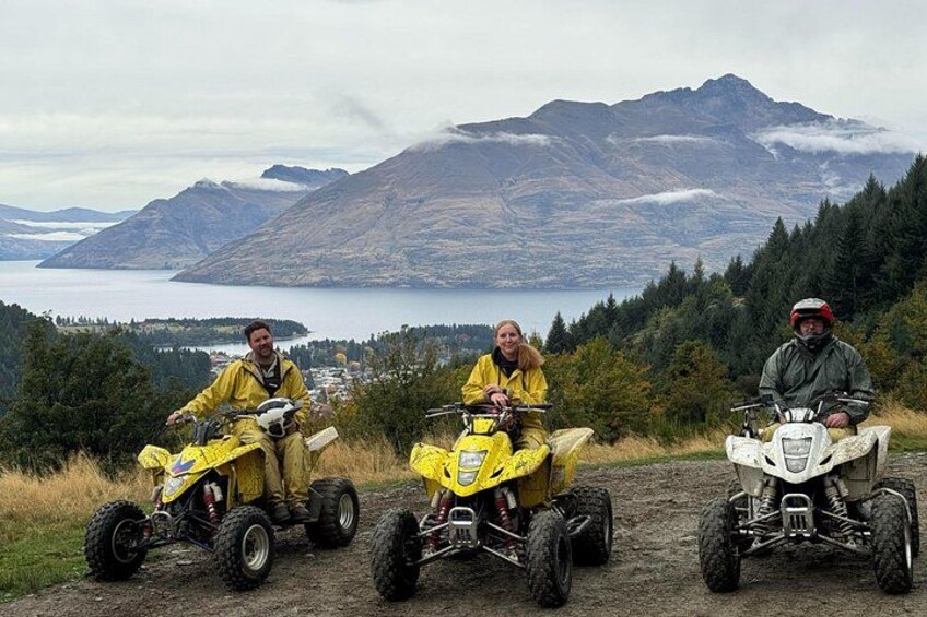 Tres personas en vehículos todo terreno (ATV) en un sendero de tierra, con un paisaje montañoso y un lago al fondo. 5