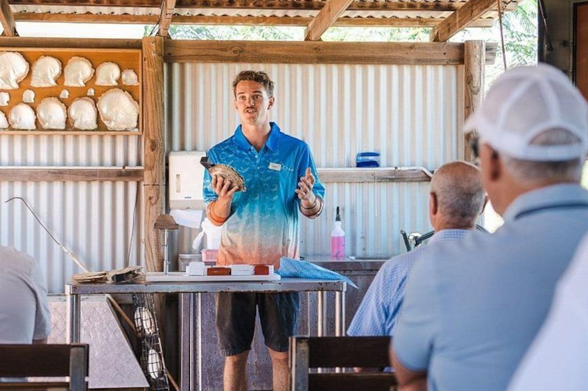 A person in a blue shirt presenting to an audience in a rustic outdoor setting with wooden structures and a corrugated metal wall. 2