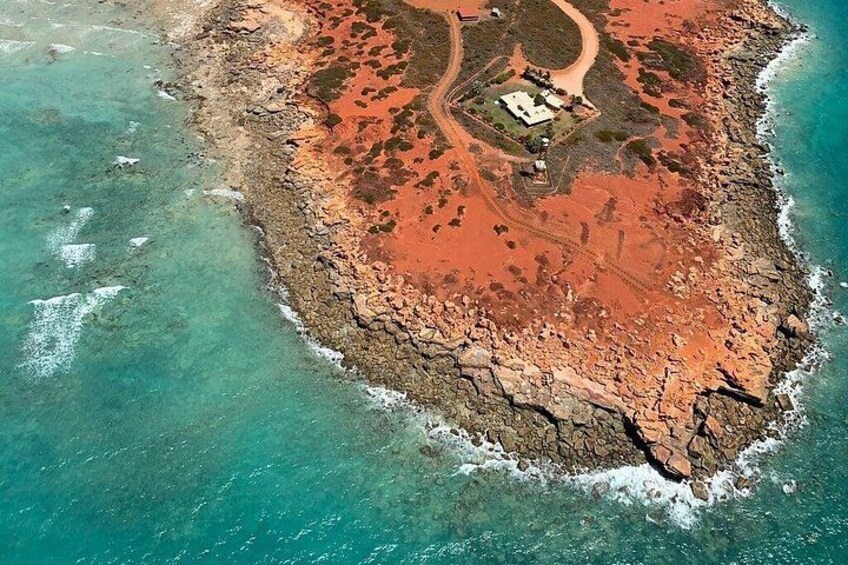 An aerial view of a coastal island with clear turquoise waters, a rocky shoreline, and a small building surrounded by a path. 4