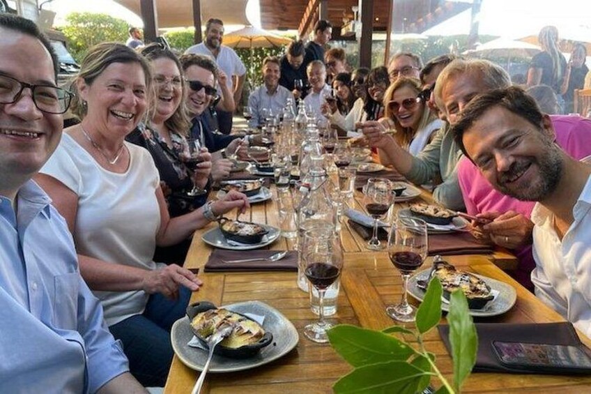 En grupp människor sitter runt ett långt träbord med vinglas och tallrikar fyllda med mat. 5