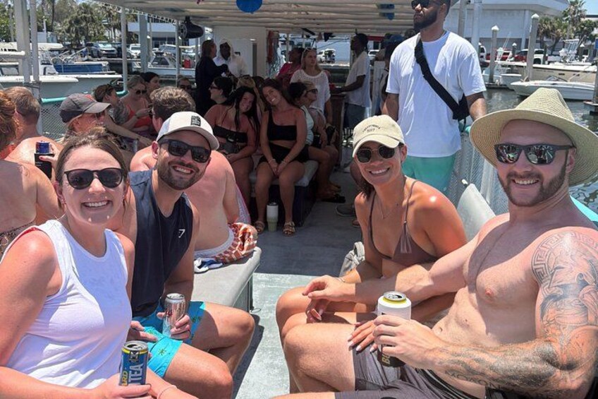 Un grupo de personas en un bote, algunas sosteniendo bebidas, con un puerto deportivo y otros botes al fondo. 5