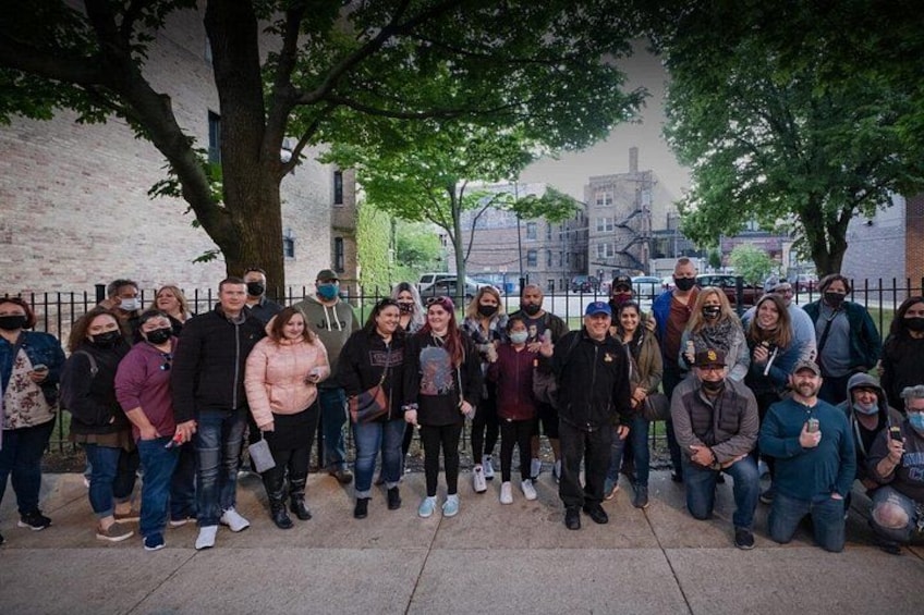 A group of people standing outdoors, some wearing masks, with trees and a brick building in the background. 2