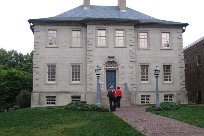 A large, historic building with a symmetrical facade, a central entrance, and a brick pathway leading to it. 1