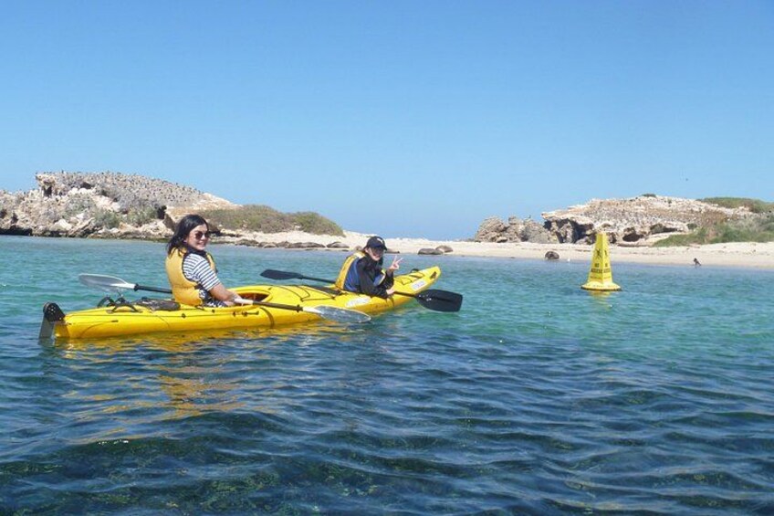 Two people kayaking on a calm sea with rocky shores and a buoy in the distance. 3