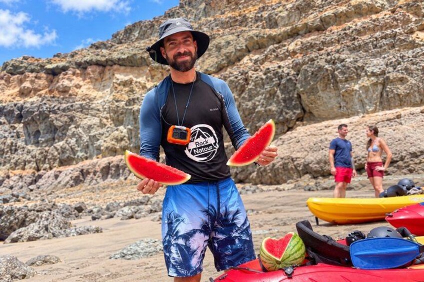 A person holding a watermelon, wearing a hat and a shirt with a logo, standing on a rocky beach with kayaks in the background. 5