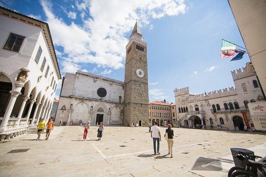 A historic square with a prominent clock tower, a fountain, and a flag. 5