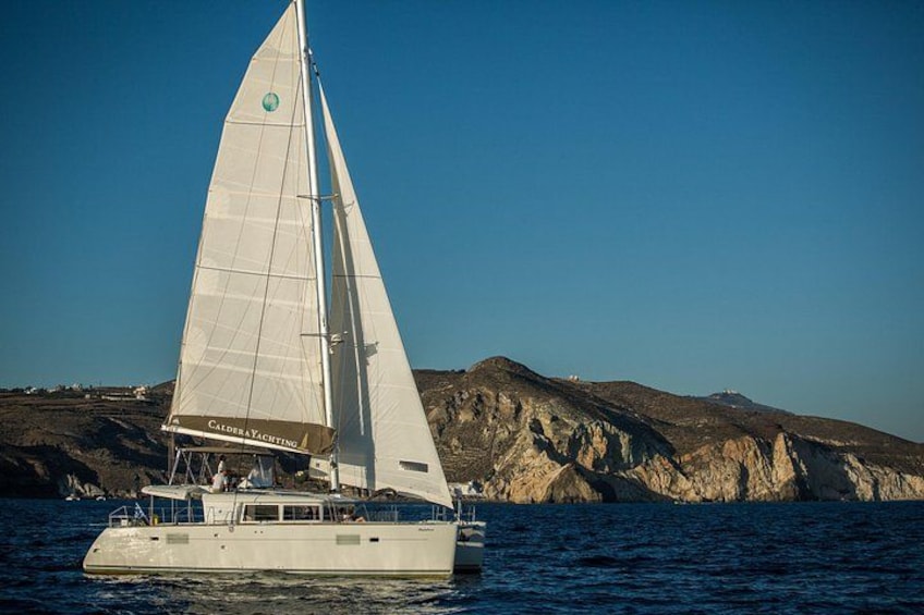 A sailboat with a white sail on calm water, with rocky hills in the background. 1
