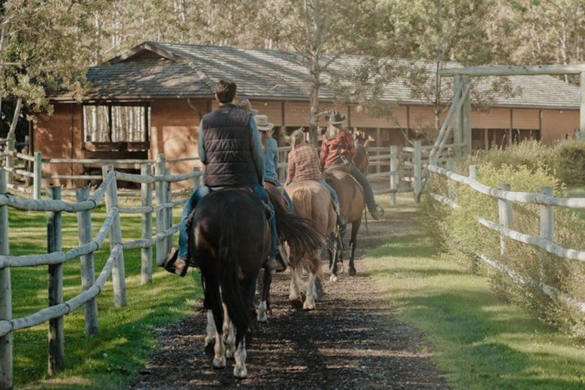 A trail with horses and riders, surrounded by wooden fences and a building in the background. 2