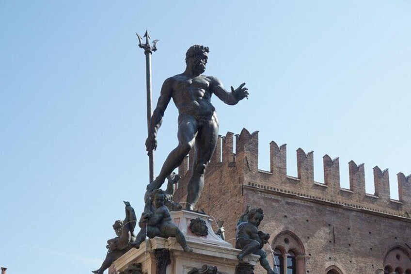 Una estatua de una figura musculosa con una lanza, sobre un pedestal con otras figuras, frente a un edificio de ladrillo con almenas. 3
