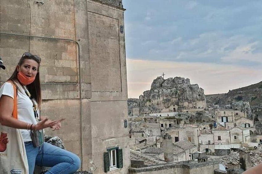 A person standing on a ledge overlooking a historic town with ancient ruins and a prominent rock formation. 3