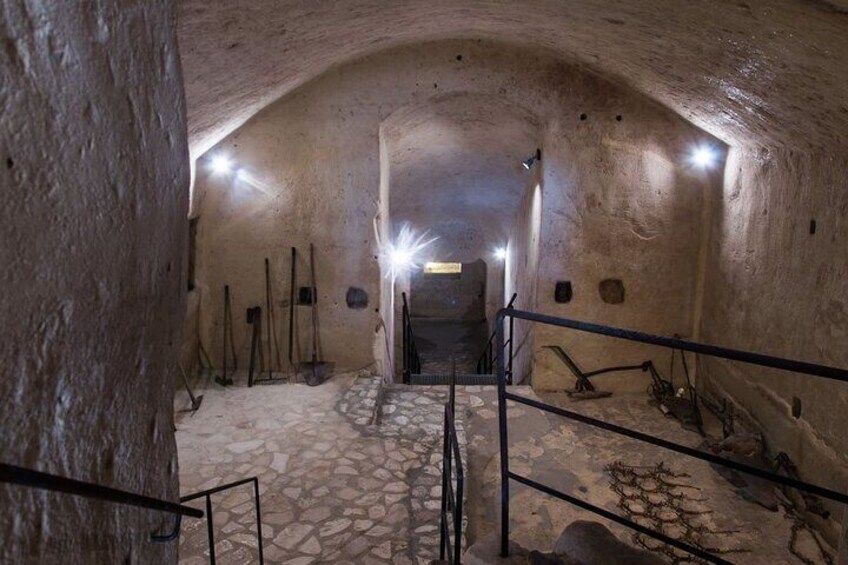 A stone-walled underground passage with a stone floor, metal railings, and a central light source. 5