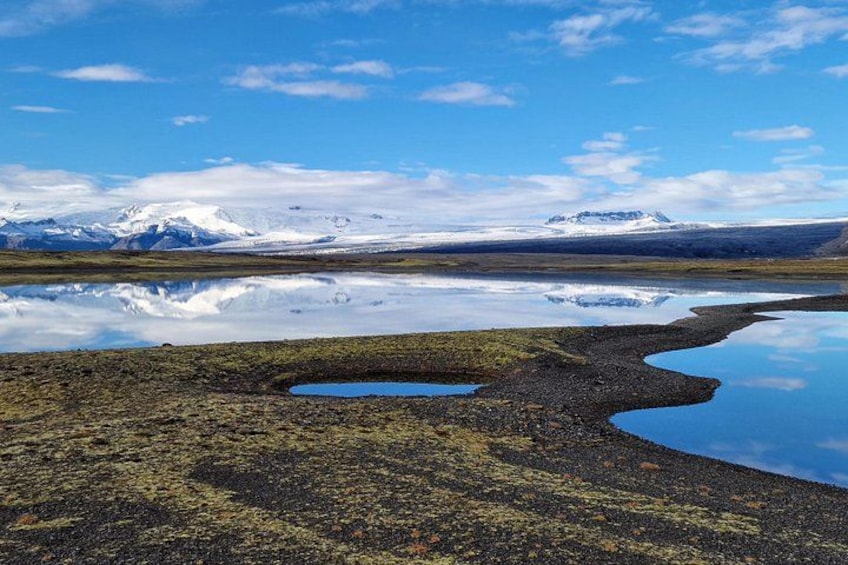 Un paisaje con una masa de agua reflectante, un primer plano rocoso y montañas distantes cubiertas de nieve. 2