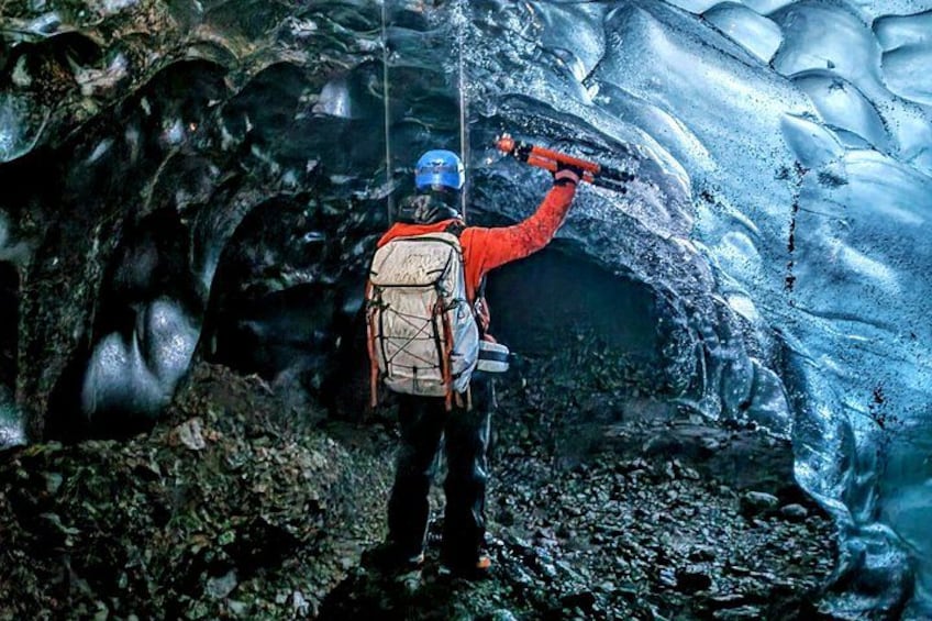 A person in a red jacket and helmet using a tool on a glacier. 2