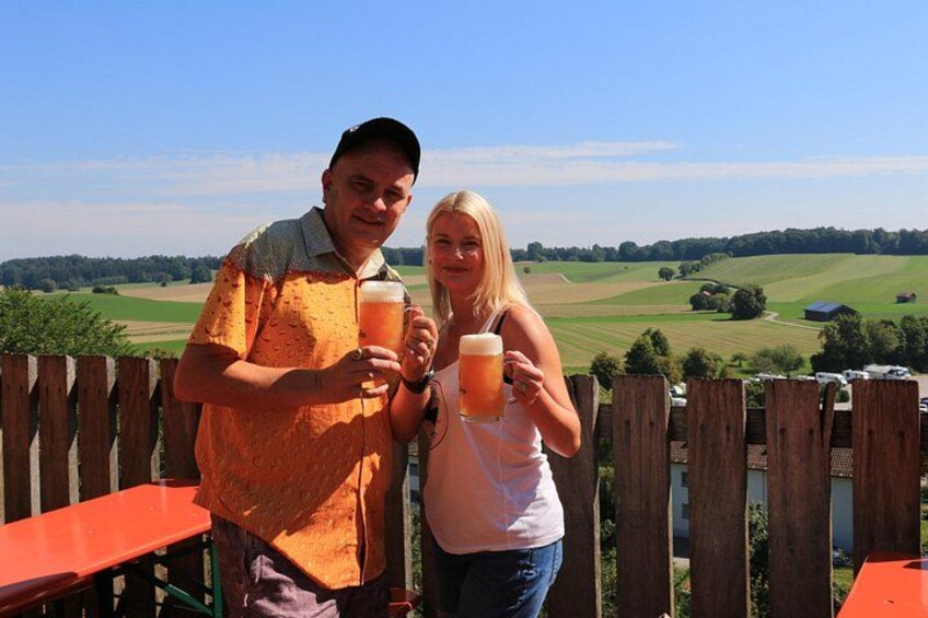 Deux personnes tenant des verres de bière, une clôture en bois et un paysage rural avec des champs et des arbres. 4