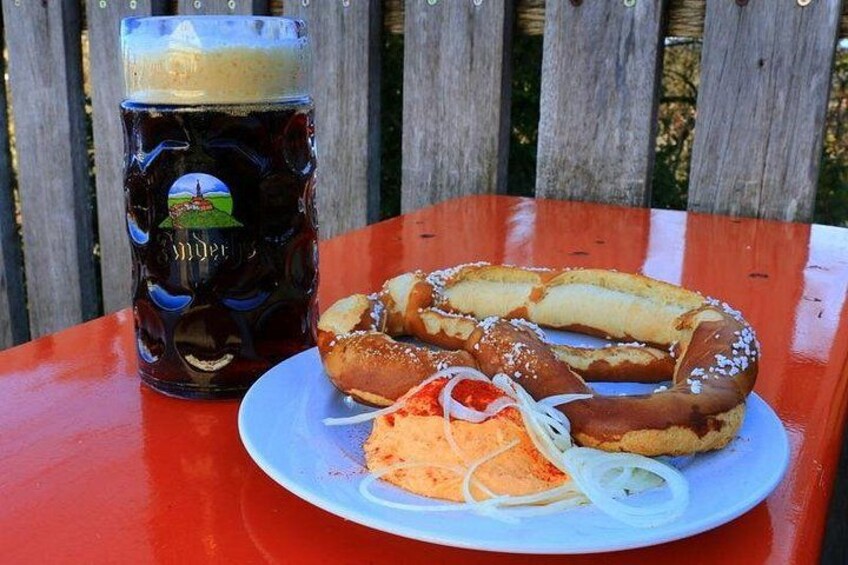 Une bière brune servie dans un verre, accompagnée de bretzels et d’oignons frits, le tout posé sur une table rouge. 3