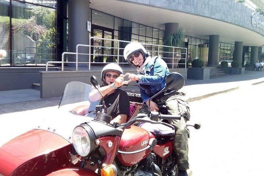 Two people on a vintage motorcycle with a sidecar, parked in front of a modern building. 4