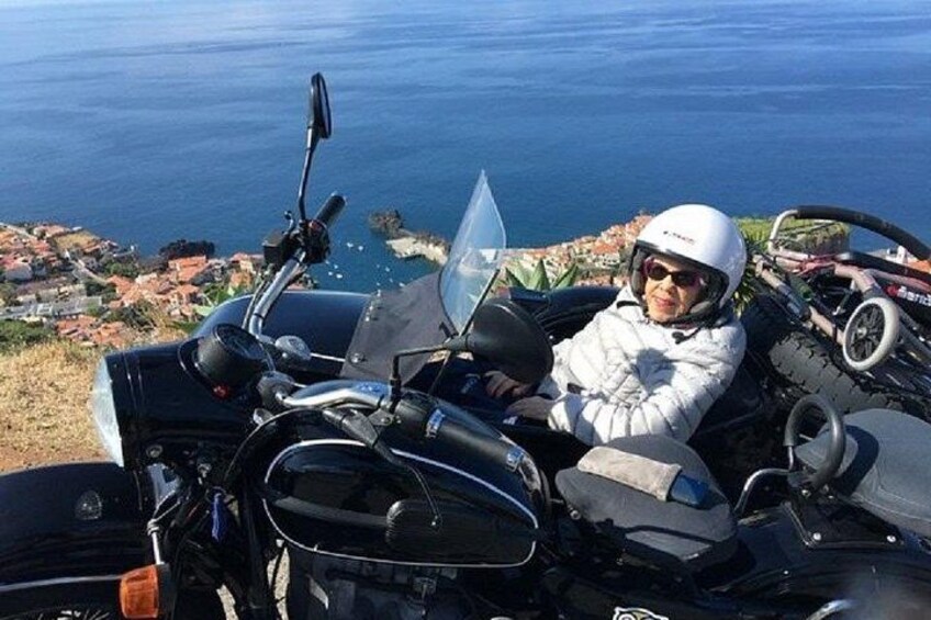 A person sitting on a motorcycle with a helmet, overlooking a coastal landscape with buildings and the sea. 3