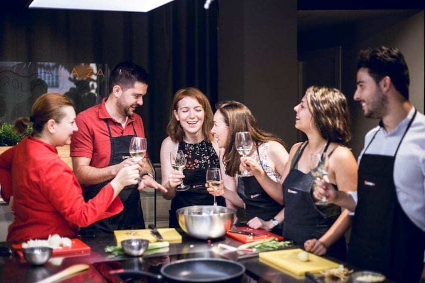 Un groupe de personnes trinquant avec des verres de vin autour d’une table où se trouvent de la nourriture et des ustensiles de cuisine. 1