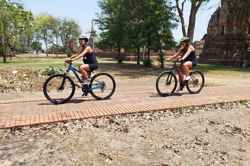 Two people riding bicycles on a paved path in a park with trees and a historical building in the background. 3