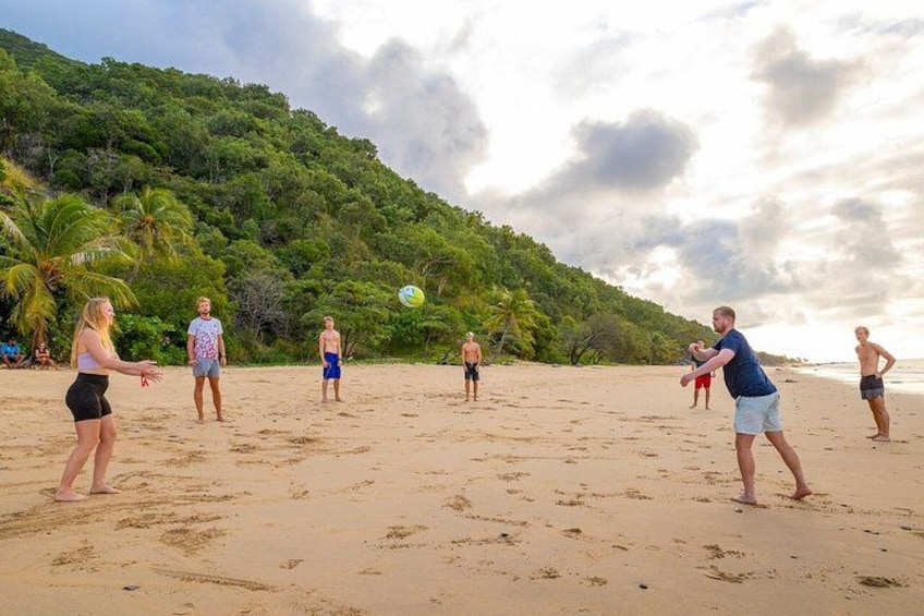 A group of people playing volleyball on a sandy beach with palm trees and a hill in the background. 2