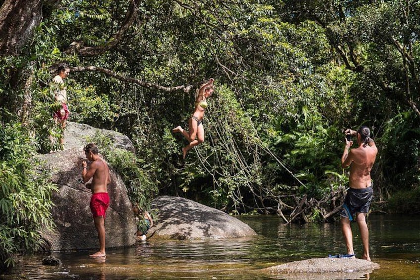 People in a forested area near a body of water, some standing on rocks and others in the water. 4