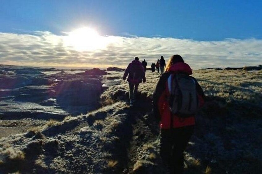 Een groep mensen wandelt over een rotsachtig, begroeid terrein, met een lage zon aan de horizon. 1