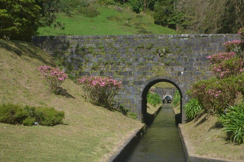 Un puente de piedra sobre un estrecho canal, rodeado de follaje y plantas en flor. 2