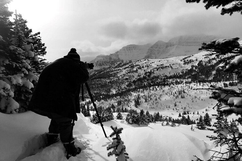 A person taking a photograph of a snowy mountain landscape with trees and a lake. 4
