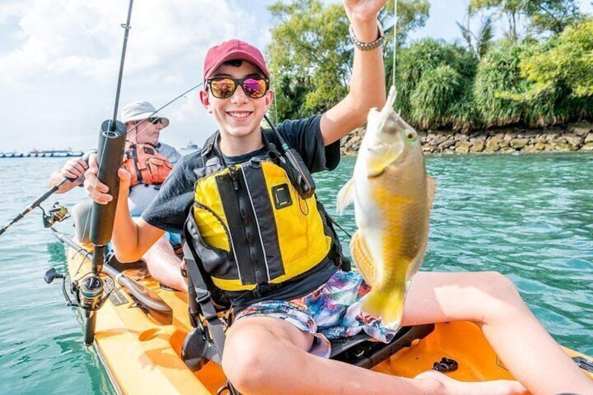 Eine Person in einem Kajak hält einen Fisch, eine andere Person fischt und im Hintergrund eine felsige Küstenlinie. 1
