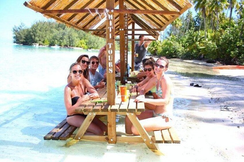 A group of people sitting at a wooden table with a thatched roof, surrounded by a beach and palm trees. 1