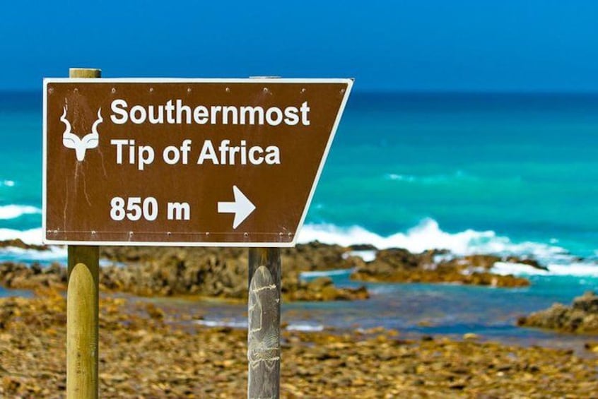 A sign indicating the Southernmost Tip of Africa at 850 meters, with a view of the ocean and rocky shore in the background. 1