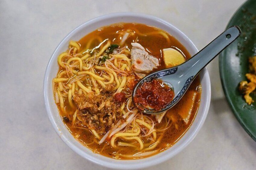 Un bol de soupe de nouilles, accompagné d’une cuillère, contenant des nouilles, de la viande et une sauce rouge. 4