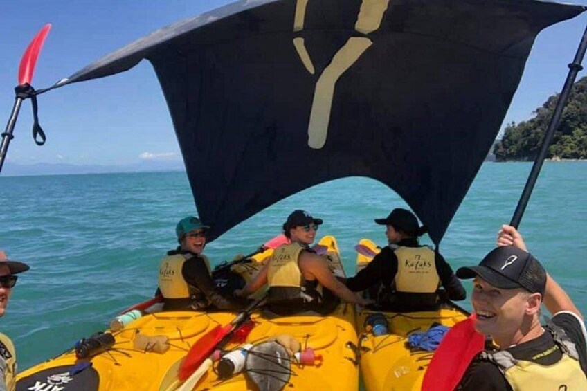 A group of people on a yellow raft with a large black canopy, wearing life jackets, on a body of water. 4