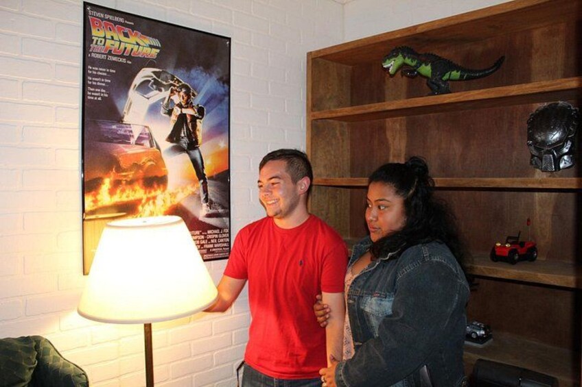 A man and a woman standing in a room with a movie poster on the wall, a lamp, and a shelf with dinosaur toys. 5
