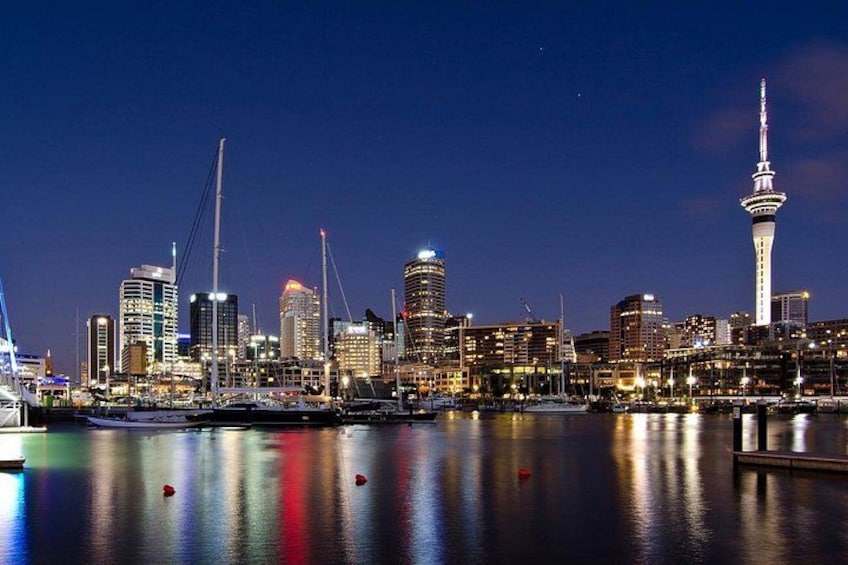 A city skyline at night with illuminated buildings, a prominent tower, and a calm waterfront. 1