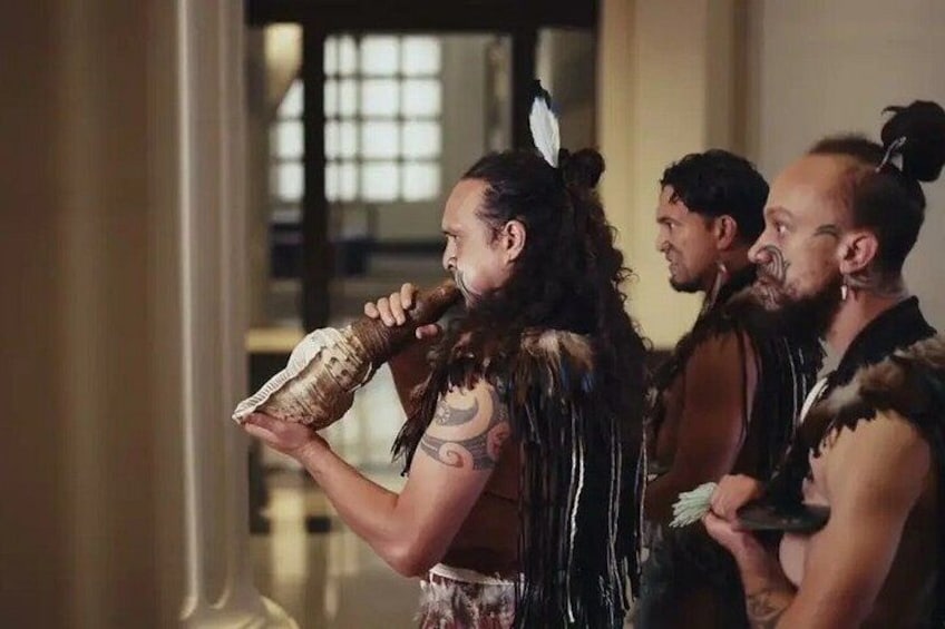 A person holding a horn-like object, wearing a feathered headdress, and another person in the background. 2